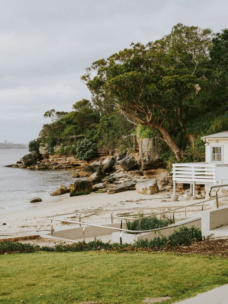 A white boat shed above a rocky beach framed by trees on the Sydney shore